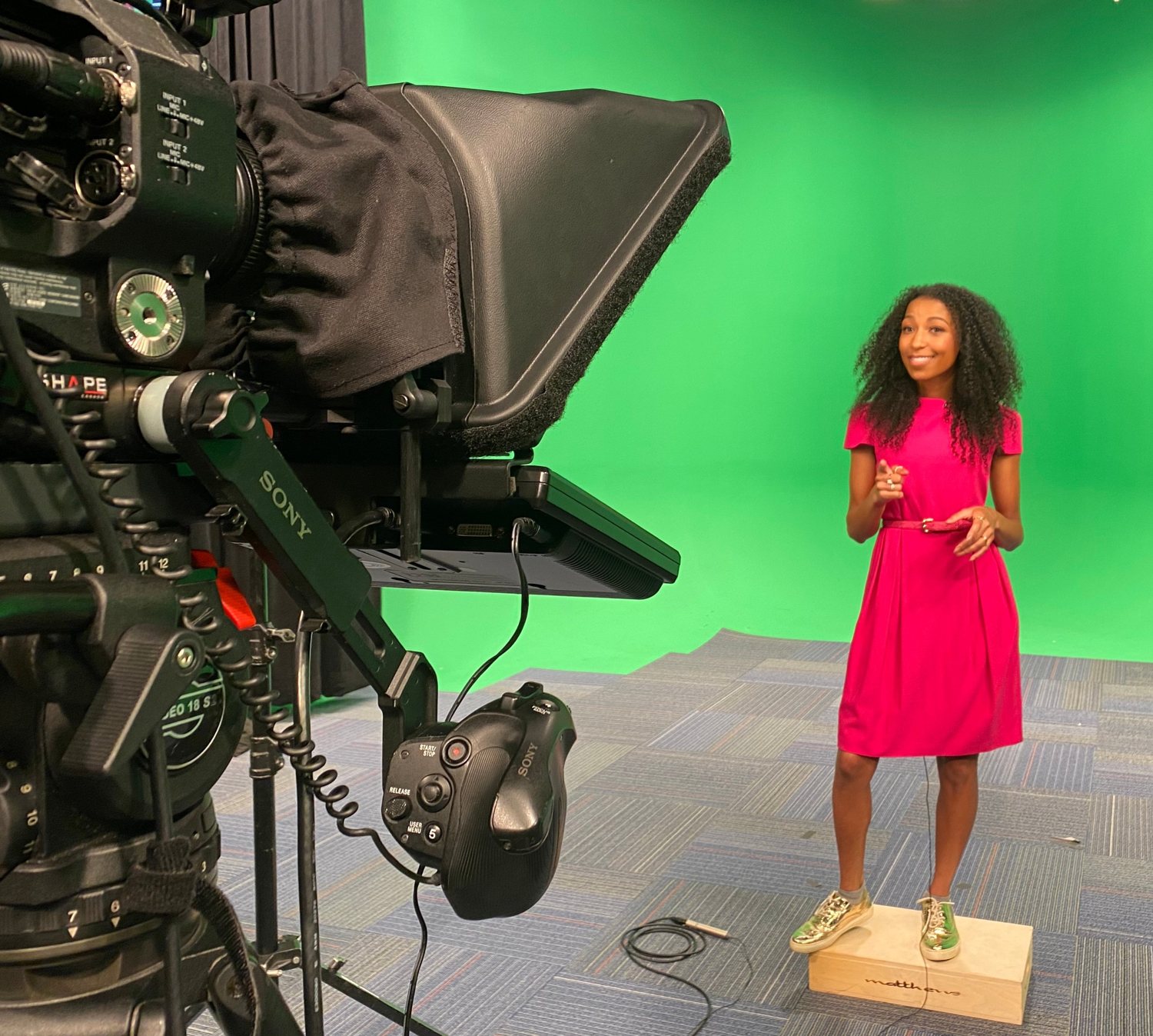 Chelsea in a pink dress delivering to a studio camera fitted with a teleprompter on a green-screen set